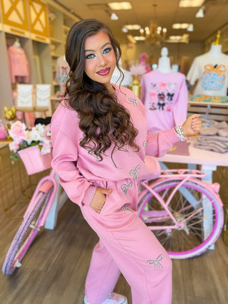 This cozy two-piece lounge set features sparkling rhinestone bows, a relaxed sweatshirt, and wide-leg pants styled with pink sneakers. Surrounded by pink boutique décor, floral accents, and feminine displays for a chic, girly aesthetic.