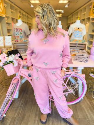 This cozy two-piece lounge set features sparkling rhinestone bows, a relaxed sweatshirt, and wide-leg pants styled with pink sneakers. Surrounded by pink boutique décor, floral accents, and feminine displays for a chic, girly aesthetic.
