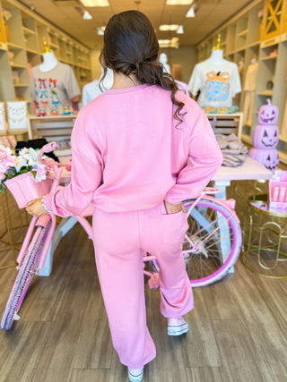 This cozy two-piece lounge set features sparkling rhinestone bows, a relaxed sweatshirt, and wide-leg pants styled with pink sneakers. Surrounded by pink boutique décor, floral accents, and feminine displays for a chic, girly aesthetic.