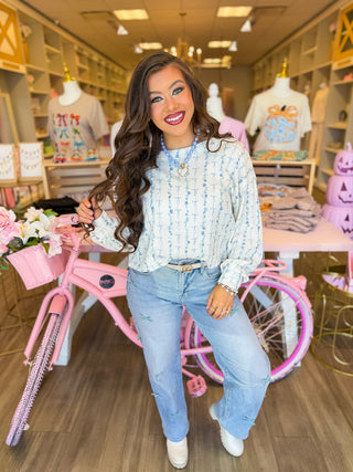 Close-up of a woman wearing a cream and blue floral waffle-knit top from Bailey Kate Boutique. The long-sleeve shirt features delicate blue flower and bow details, styled with denim jeans, gold bracelets, and a blue beaded heart necklace for a soft, feminine look.