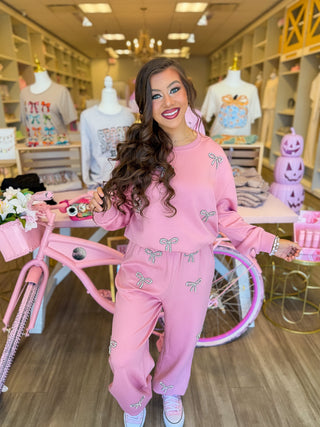 This cozy two-piece lounge set features sparkling rhinestone bows, a relaxed sweatshirt, and wide-leg pants styled with pink sneakers. Surrounded by pink boutique décor, floral accents, and feminine displays for a chic, girly aesthetic.