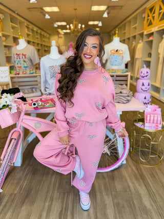 This cozy two-piece lounge set features sparkling rhinestone bows, a relaxed sweatshirt, and wide-leg pants styled with pink sneakers. Surrounded by pink boutique décor, floral accents, and feminine displays for a chic, girly aesthetic.