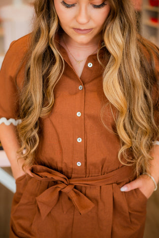 Woman wearing a rust-brown button-front belted romper with cream ric-rac trim on sleeves and shorts hem, featuring short sleeves, side pockets, and a waist tie belt, standing in a boutique setting.