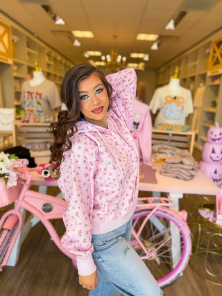
Woman modeling a pink floral hoodie inside Bailey Kate Boutique in Somerset, Kentucky. The soft pastel sweatshirt features a dainty rose print, drawstring hood, and relaxed fit, styled with light denim bow jeans for a cozy, feminine boutique look surrounded by girly pink décor and displays.
